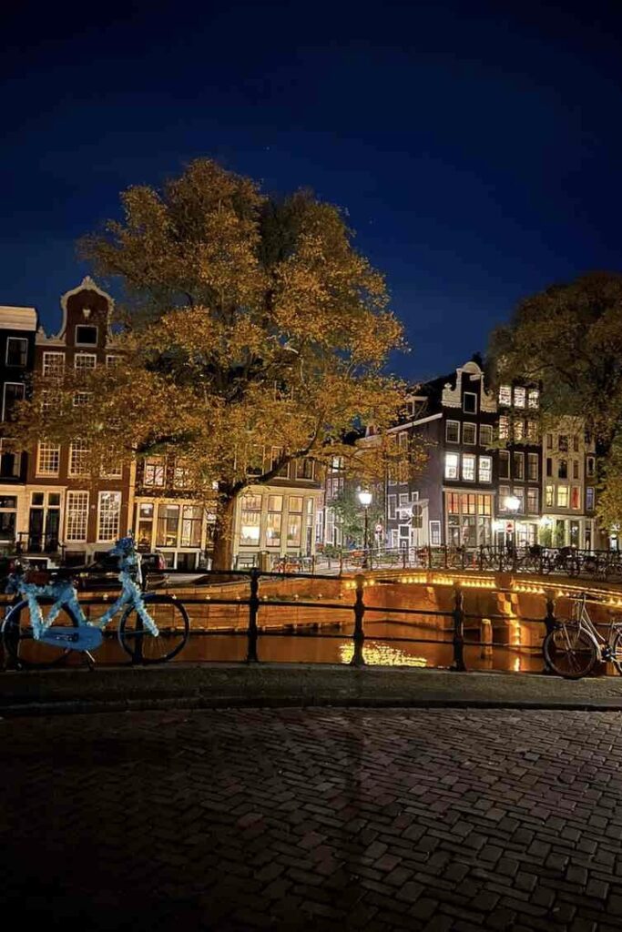 Scenic view of Amsterdam canal at night with illuminated bridge, traditional Dutch houses, and bicycles along the water under a clear dark blue sky.