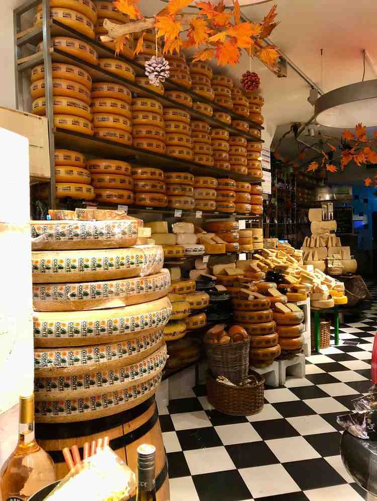 Traditional Dutch cheese shop in Amsterdam with shelves stacked with large wheels of Gouda and Edam cheese, autumn decorations, and checkered black-and-white floor tiles.